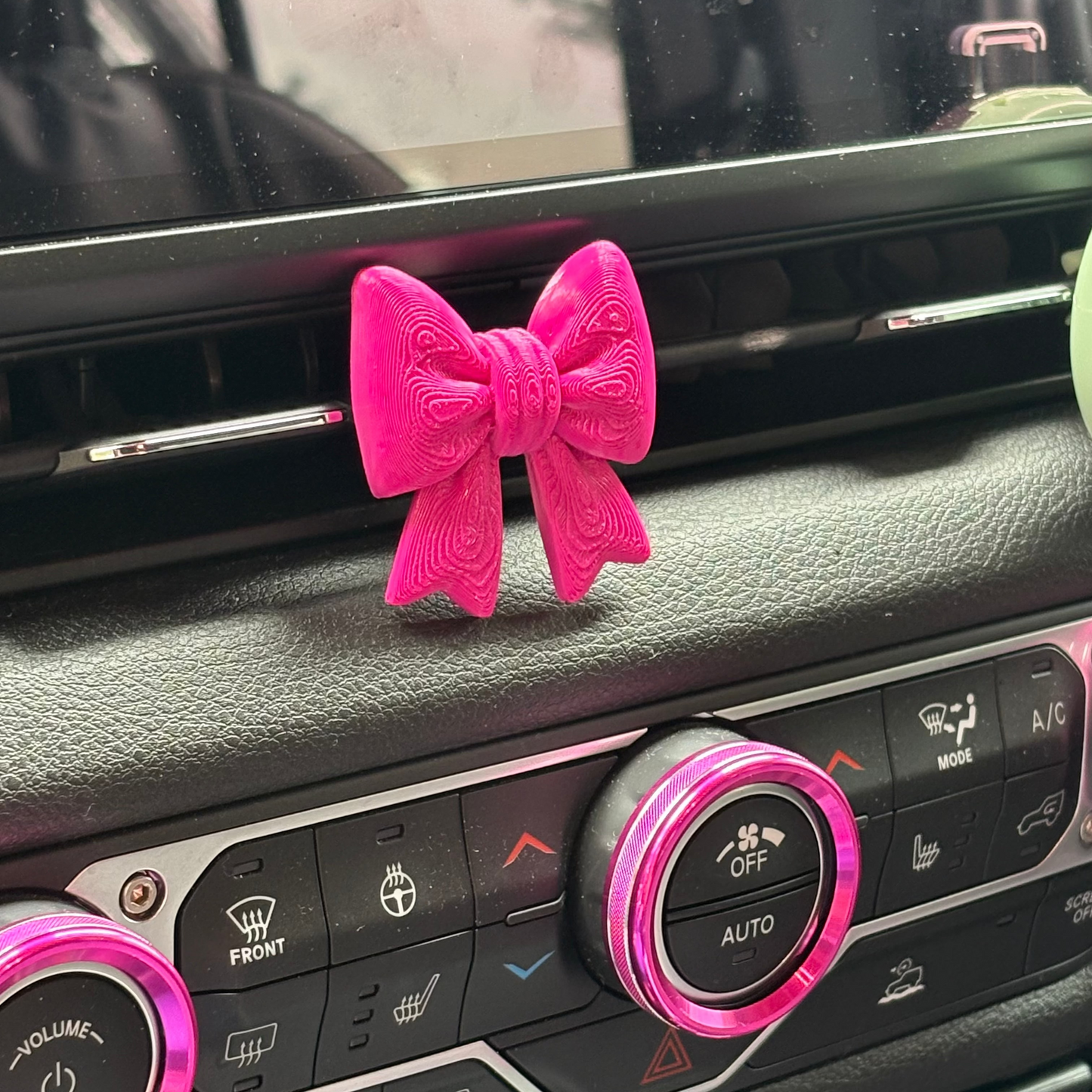 A bright pink bow-shaped car air freshener or dashboard accessory placed on the vent of a vehicle's climate control console.