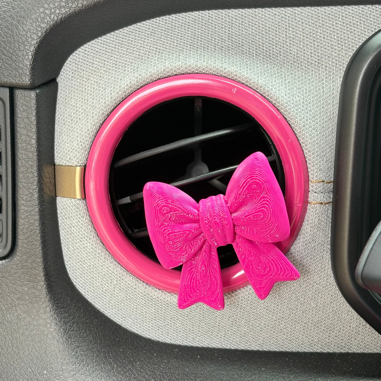 A hot pink clip shaped like a bow, shown inserted into a circular car air vent.